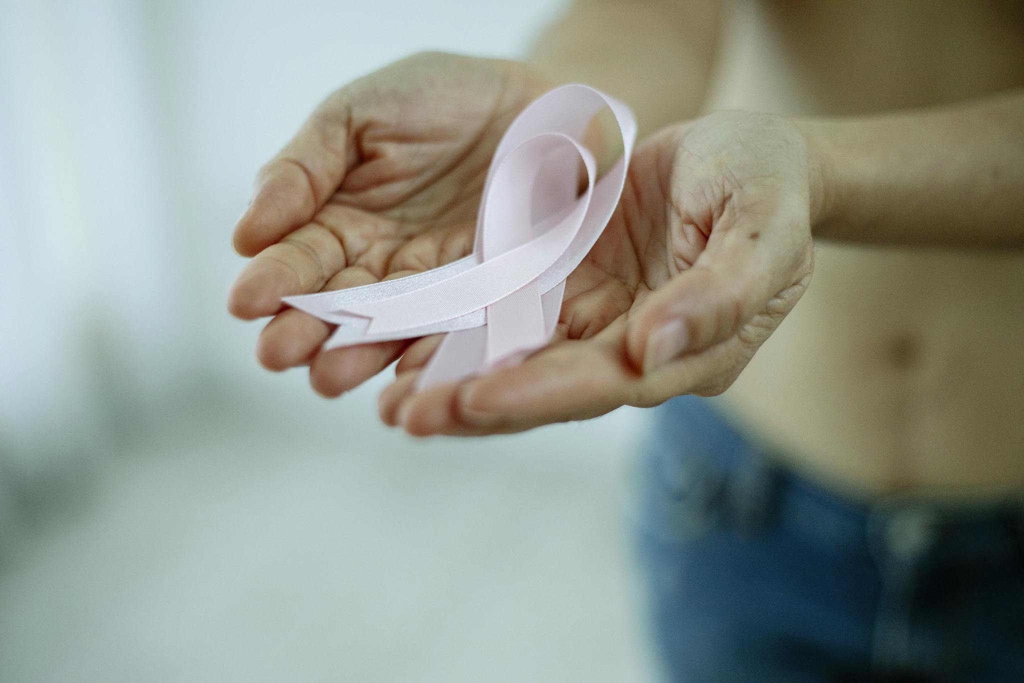 Crop unrecognizable topless lady demonstrating pink ribbon in arms as symbol of breast cancer awareness while standing in bright room